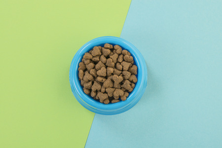 Blue round feeding bowl with dark brown heart shaped pet kibble on a blue and soft green background