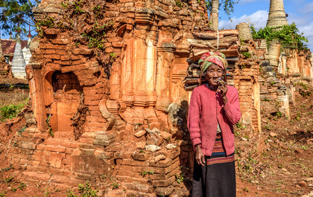AVA, MYANMAR - JANUARY 27, 2016 : old wrinkled woman smokes  a cheroot cigar around ruined temples in the ancient city  of Ava, also called Inwaのeditorial素材