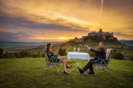 Couple in love drink red wine in nature under the ruins of a castle at sunset. Boyfriend trows a tulip to his girlfriend.の写真素材