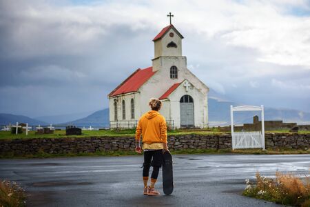 Young man with a skateboard looking at a church with a cemetery in Icelandの写真素材
