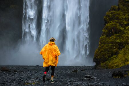 Tourist wearing a yellow raincoat walks to the Skogafoss waterfall in Iceland