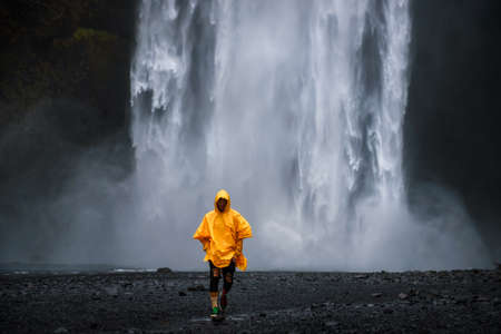 Tourist wearing a yellow raincoat walks from the Skogafoss waterfall in Icelandの写真素材