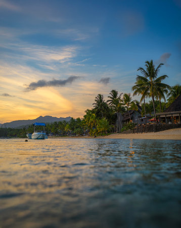 Serene beach in Bel Ombre, Mauritius, at sunset with calm waters, a boat floating near the shore, palm trees, beachfront resort huts, and distant mountains under a colorful sky.の写真素材