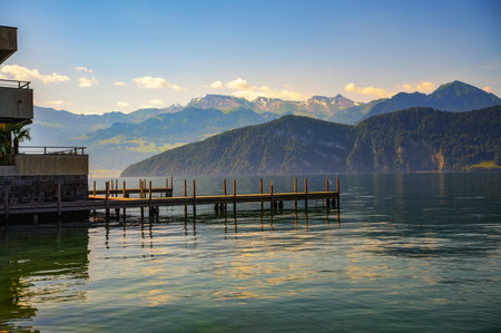A peaceful sunset over a wooden jetty on Lake Lucerne in Weggis, Switzerland, with reflections on the water and the Swiss Alps in the background.の写真素材
