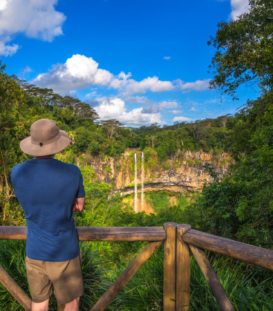 A tourist wearing a hat and casual clothing stands at a wooden viewpoint, looking at a waterfall surrounded by lush greenery.の写真素材