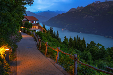 Evening view of Lake Brienz and the Harder Kulm area with a lit walking path and surrounding Swiss Alps under dramatic clouds in Switzerland.の写真素材