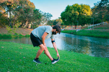 A young man doing stretching in a green park before starting to exercise in sportswear and mask.