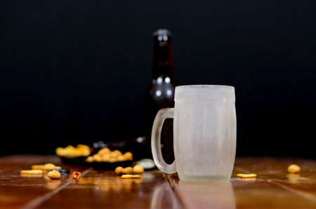 A cold empty beer mug with two beer bottles behind with some savory snacks on a wooden table with a...