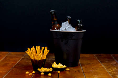 Black ice bucket with four beer bottles inside on a wooden table and with some savory snacks with a...