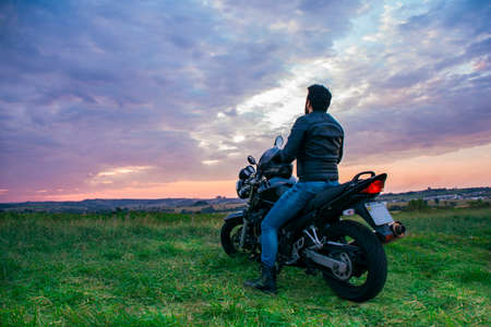 Man sitting on a black motorcycle, wearing blue jeans, a black jacket and a black helmet, with his back turned against a landscape in the background.の写真素材