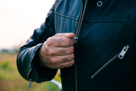 Man whose face is not visible, dressed in jeans and a black jacket, zipping his jacket with a motorcycle behind him on the street.の写真素材