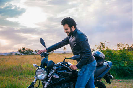 Man standing next to a motorcycle, wearing jeans and a black jacket, looking at the motorcycle with...