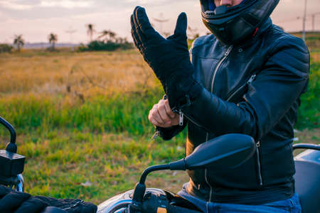 Man sitting on a black motorcycle, dressed in jeans and a black jacket, putting on black gloves with the landscape in the background.の写真素材