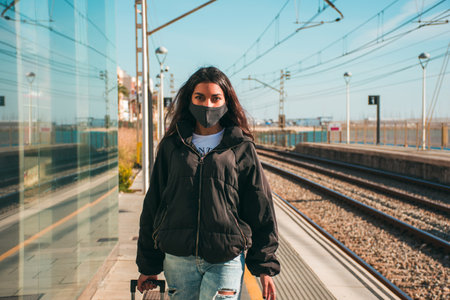 A young woman with brown skin with a black face mask walking beside the train tracks in the middle...
