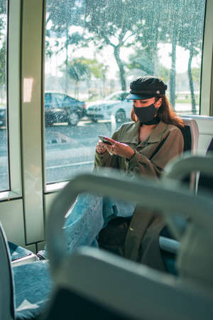 A young woman with brown skin wearing a black protective face mask sits inside public transport using her mobile phone.の写真素材