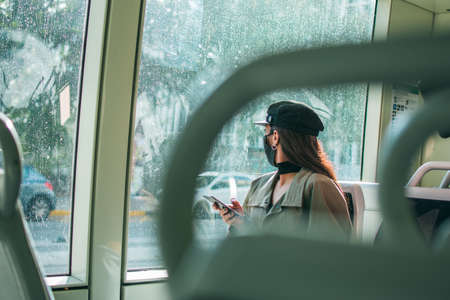 A young woman with brown skin wearing a black protective face mask sits inside public transport...