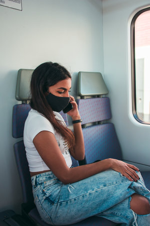 A young woman with brown skin wearing a black protective face mask using the mobile phone in public transport.の写真素材
