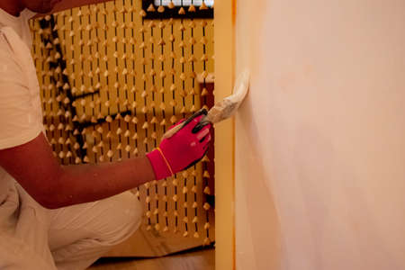 Dark-haired man with a dark beard painting a cream-colored wall with white paint with a brush to...