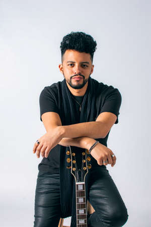 Portrait of a Latin man with dark skin and beard, dressed in black leaning on a red electric guitar...