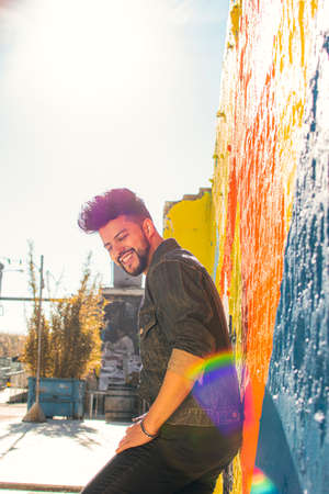 Urban photography in which a brown-skinned Latino man with dark hair and appears beard, dressed in a denim jacket, leaning against a graffiti wall laughing.の写真素材