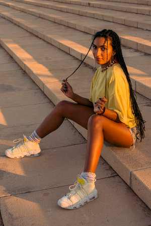 Pretty young woman with dark skin with long black braids sitting in warm colored summer clothes on a concrete stairs on the seafront at summer sunset.の写真素材