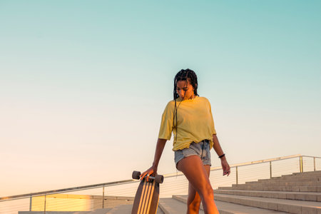 Pretty young woman with brown skin with long black braids standing holding a skateboard with her hand dressed in warm colored summer clothes on a concrete stairs on the seafront at summer sunset with clear blue sky.の写真素材