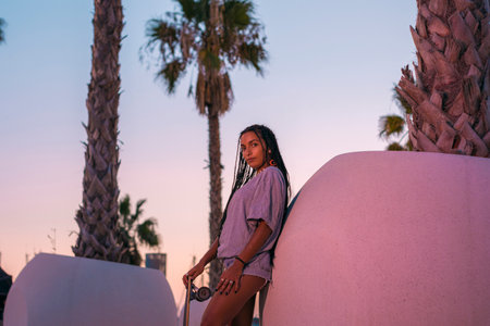 Pretty young woman with brown skin with long black braids posing with her skateboard dressed in a...