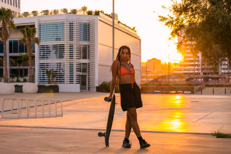 Pretty young woman with brown skin with long black braids posing with her skateboard dressed in an orange bikini and black shorts having fun on the seafront in the summer sunset in the warm light with palm trees in the background.の写真素材