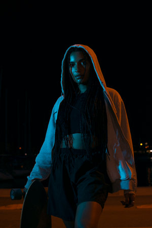 Pretty young woman with brown skin with long black braids holding her skateboard dressed in a gray...