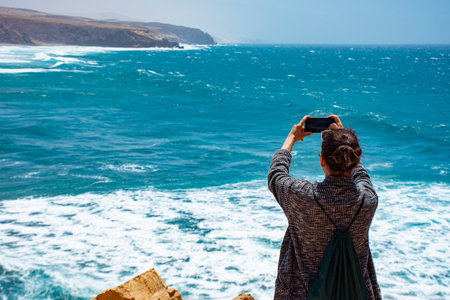 A white mature woman on some rocks next to the sea taking photographs with her smartphone of the...