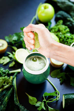 A woman is squeezing a lemon on a green juice of healthy, fresh and raw vegetables on a black background. Kiwi, kale, lemons, green apple, parsley.の写真素材
