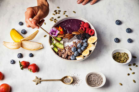 A woman is eating an aÃ§ai bowl with fresh fruits for breakfast with banana, blueberries, blackberries, strawberries, kiwi, nuts, and seeds.の写真素材