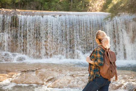 A girl watches a waterfall on an autumn day. She wears a backpack and plaid shirt.の写真素材
