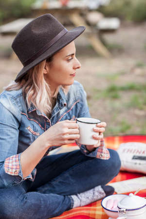 A girl enjoying a cup of hot drink outdoors having a picnic in nature. She wears a hat and denim shirt.の写真素材