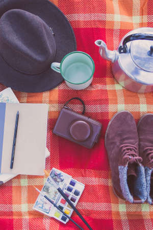 Overhead view of various artist objects: watercolors, notebook, boots, camera, hat, mug and teapot on a red picnic blanket.の写真素材
