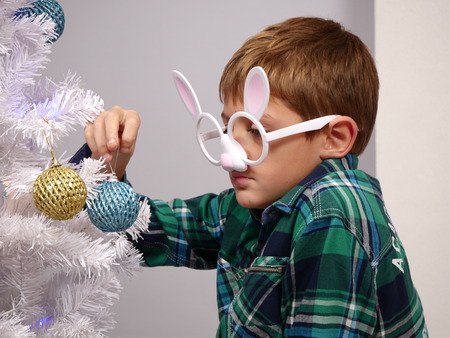 Cute boy in funny glasses decorating white Christmas tree with colorful beadsの写真素材