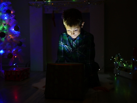 child boy opening gift box and surprises near christmas decorated fir tree, dark backgroundの写真素材