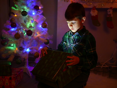 child boy opening gift box and surprises near christmas decorated fir tree, dark backgroundの写真素材
