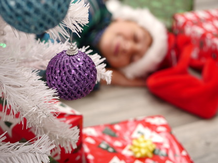 Cute boy in Christmas hat cutely sleeping on the floor behind the white decorated Christmas treeの写真素材