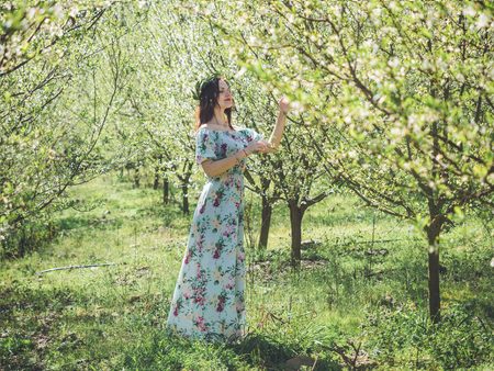 Young beautiful brunette woman in long dress in bay leaf wreath standing dreamily with closed eyes in spring blossom cherry garden.の写真素材