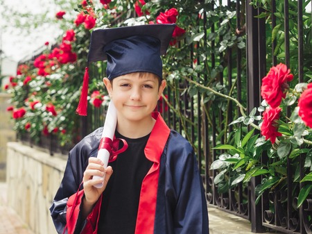 Happy caucasian child in graduation gown with diploma standing near stone fence full of wild roses. Students celebration graduation, education conceptの写真素材