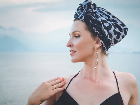 Portrait of a young, attractive Muslim woman with bright skin, red lipstick, beautiful manicure, headscarf and earrings on the ears standing still calm blue sea on backgroundの写真素材