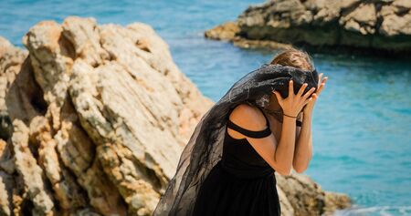 Young beautiful woman in black dress on the rocks by the sea in windy weather. Closing her face with black transparent scarf.の写真素材