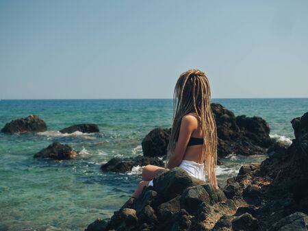 Dreadlocks fashionable girl sitting on the rocks near turquoise color Mediterranean sea. Meditating...