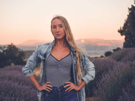 Young blonde woman in denim jacket standing in lavender field sunset time.の写真素材