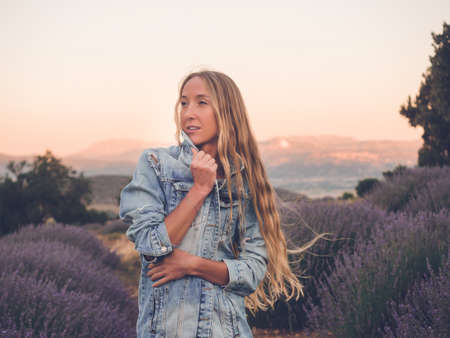 Young blonde woman in denim jacket standing in lavender field sunset time.の写真素材