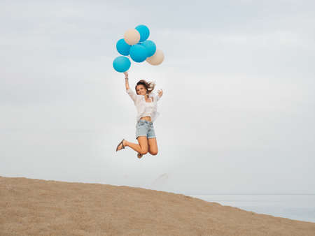 Young happy woman jumping with blue and white helium balloons on sandy beach near the sea in cloudy...