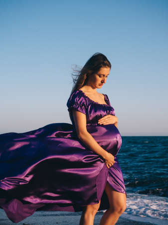 Young beautiful woman 7 month pregnant in violet dress looking at her belly on Mediterranean sea backgroundの写真素材