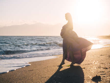 Young beautiful woman 7 month pregnant in violet dress enjoying Mediterranean sea in sunset timeの写真素材