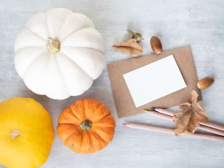 Empty white small paper on retro style envelope, some colorful pencils and autumn oak leaves, acorns and decorative pumpkins on the tableの写真素材